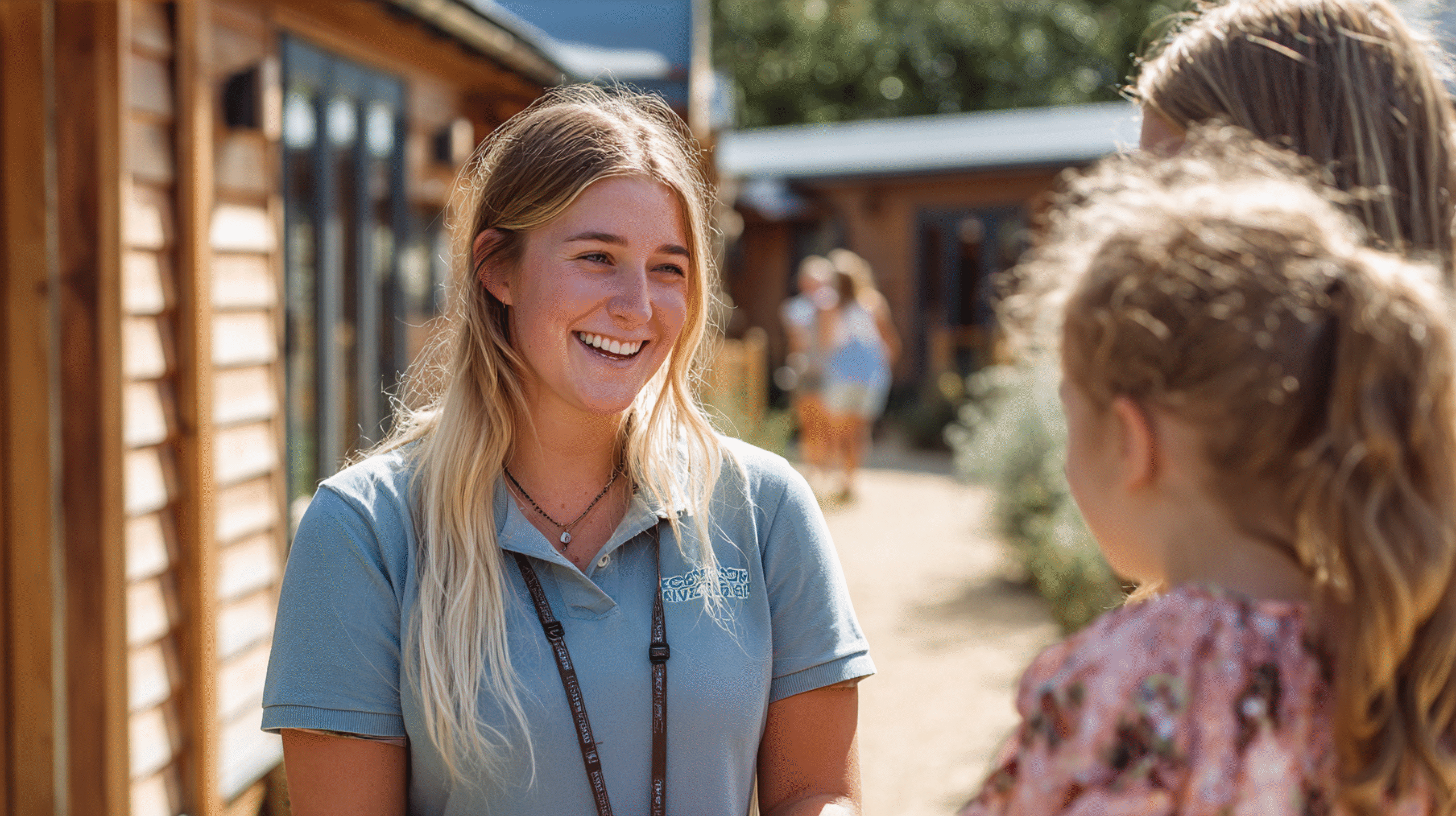 Holiday park staff member smiling and speaking to a child and adult guest outside timber lodge accommodation in bright summer light