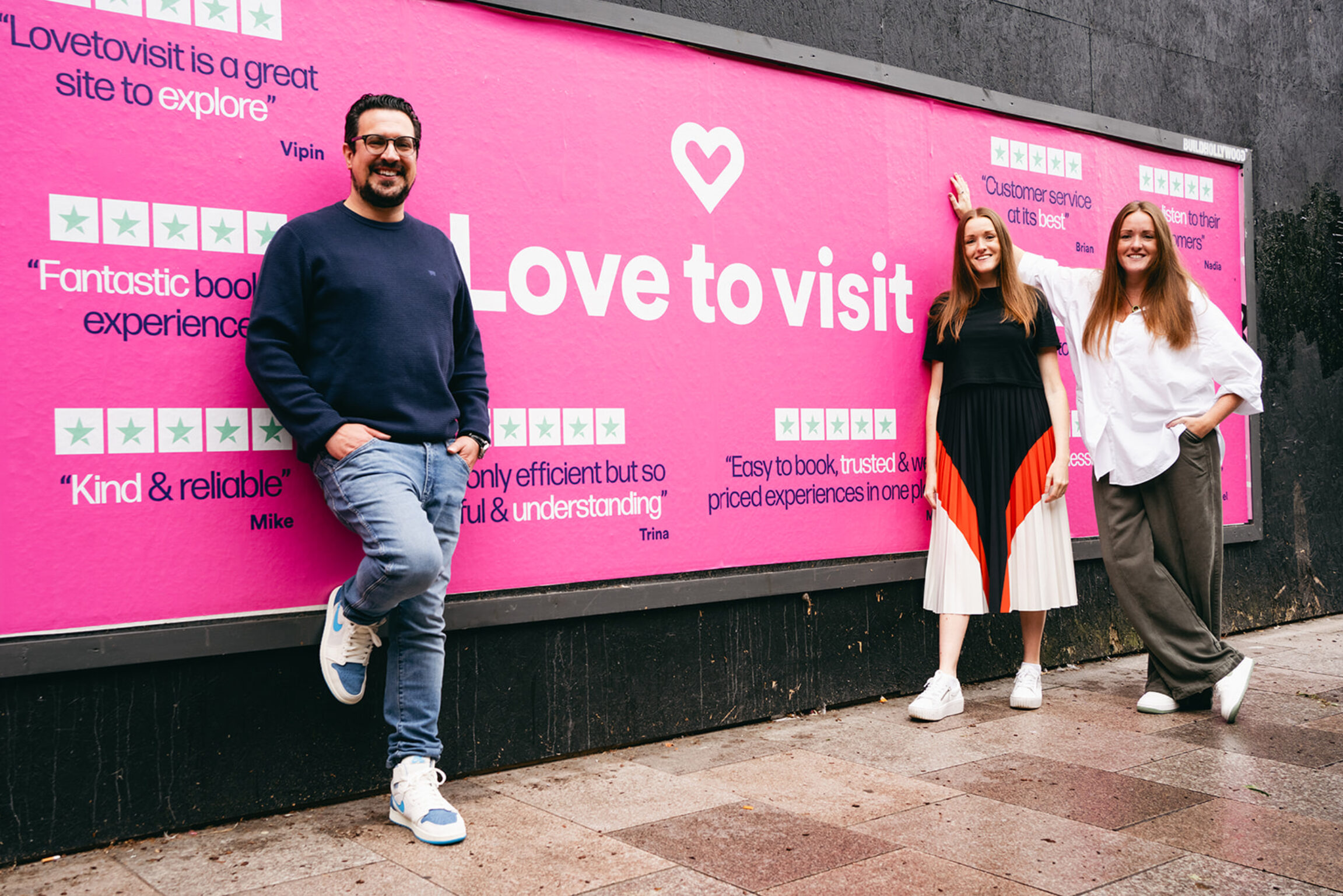 Fed Pereira with co-founders Alice and Georgia from LoveToVisit, standing in front of a bright pink LoveToVisit billboard.